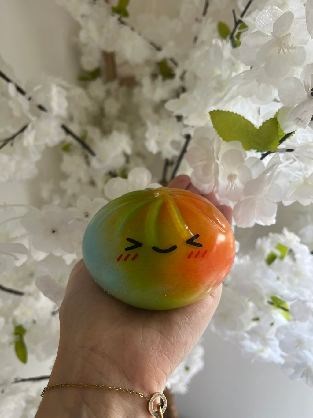 Squishy bao orange et vert dégradé tenu dans une main devant des fleurs blanches, montrant sa taille réelle